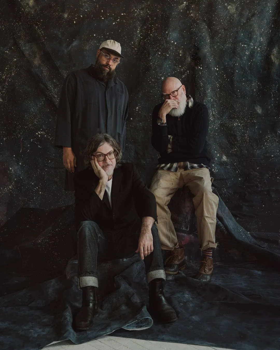 Three men pose against a dark, textured backdrop with speckled lighting; one seated wearing glasses and dark jacket, one…