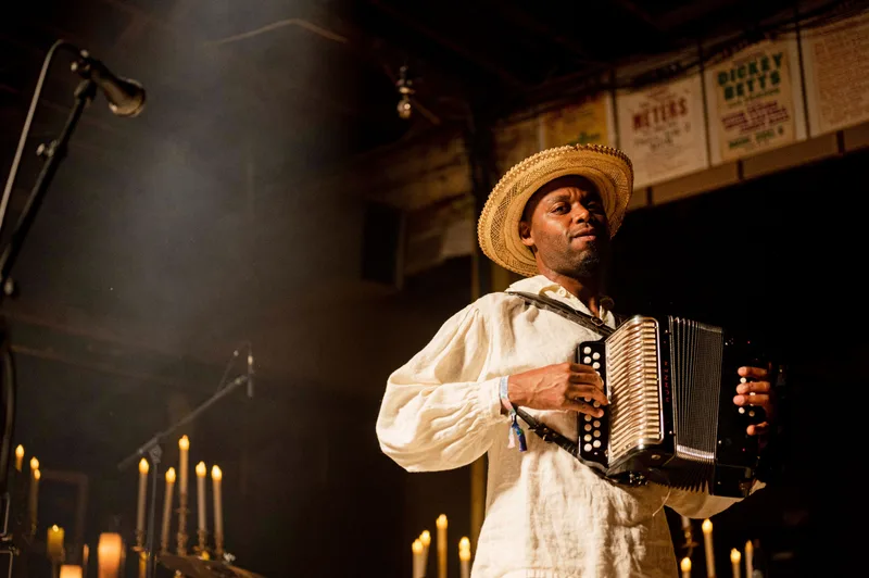 Musician playing accordion on stage wearing a cream linen shirt and straw hat, surrounded by candles and a microphone.