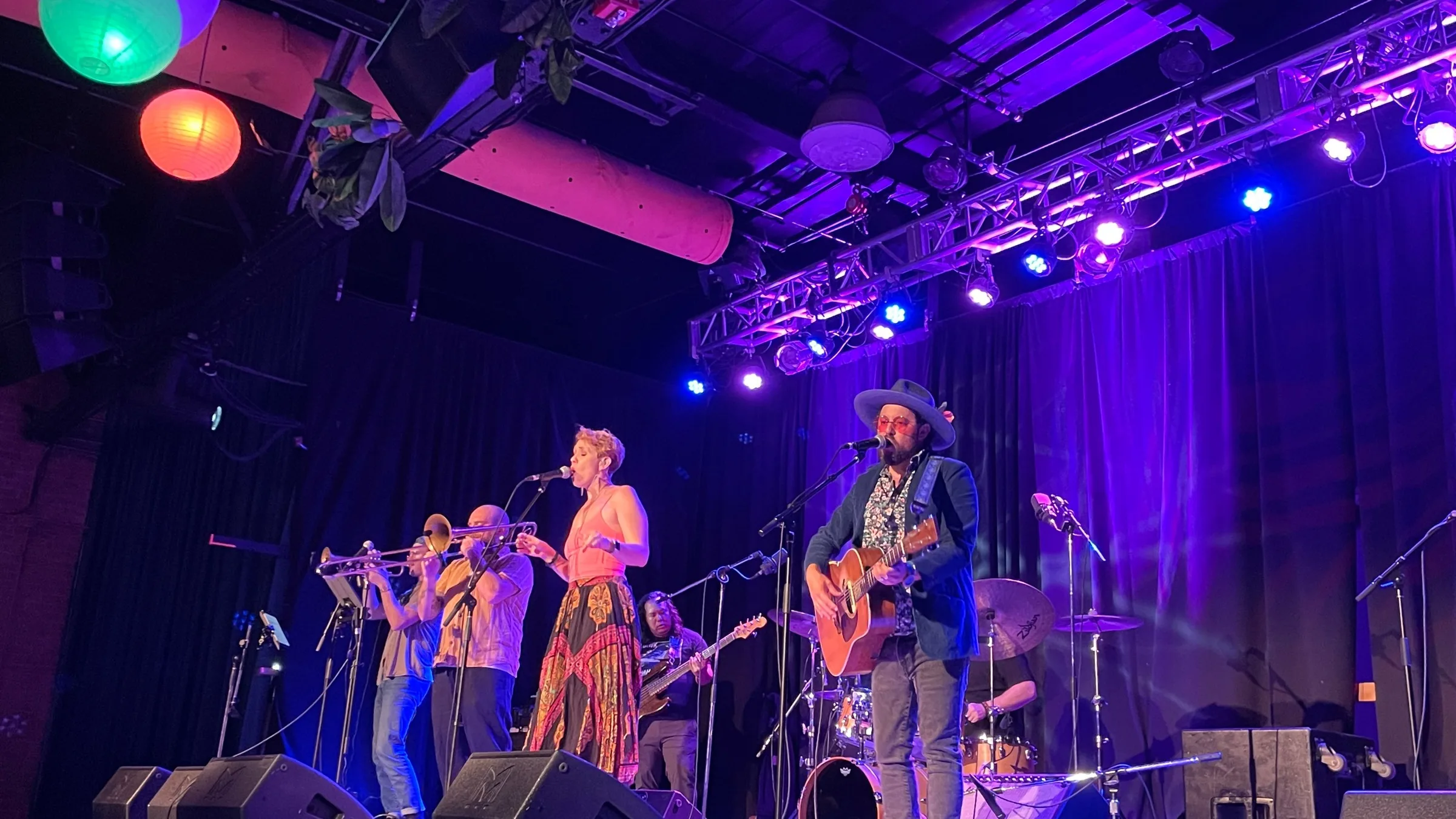 A band with trombone, trumpet and acoustic guitar on stage under violet light and green and orange lanterns