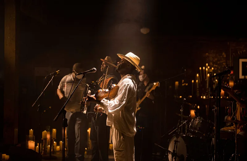 A saxophonist in a straw hat and white shirt performs center stage while a vocalist sings at a microphone, with band…