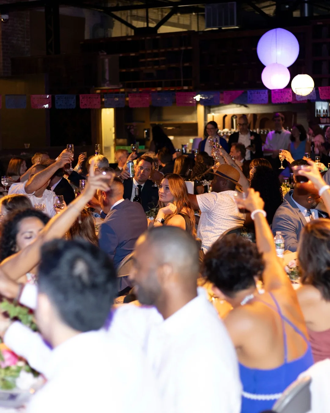 Guests at long banquet tables raising their glasses, under paper lanterns and a string of papel picado