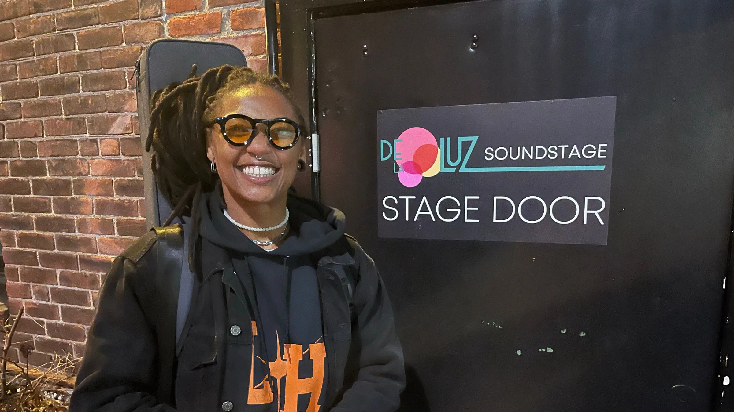 A musician in round orange-tinted glasses, a guitar case on her back, smiling beside the De la Luz Soundstage stage door