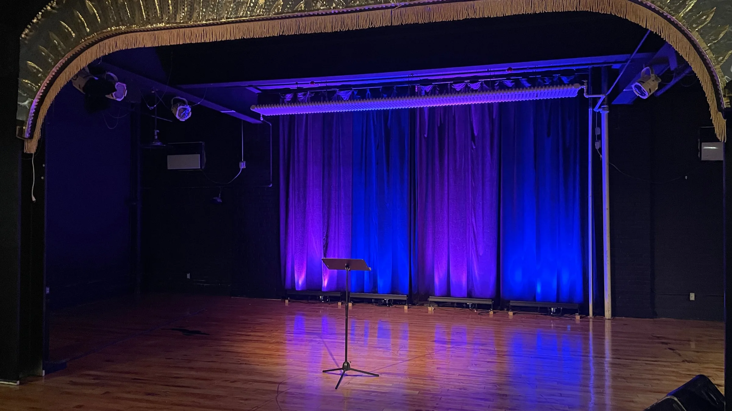 The empty Divine Theater: a gilded, fringed proscenium arch framing a wooden floor and curtains lit violet and blue
