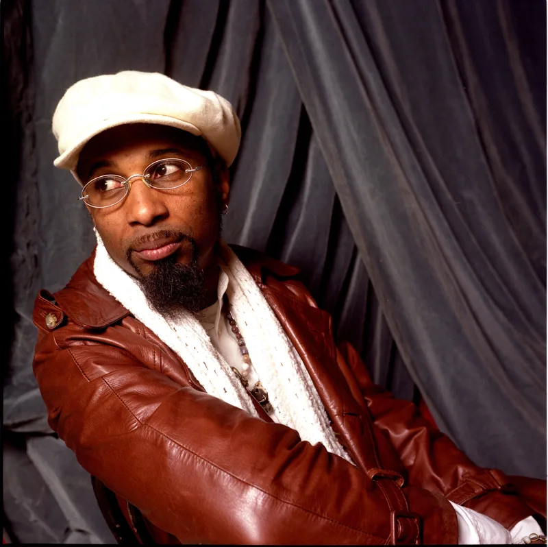 Man wearing a cream-colored cap, eyeglasses, and burgundy leather jacket over a white shirt against a dark draped backdrop.